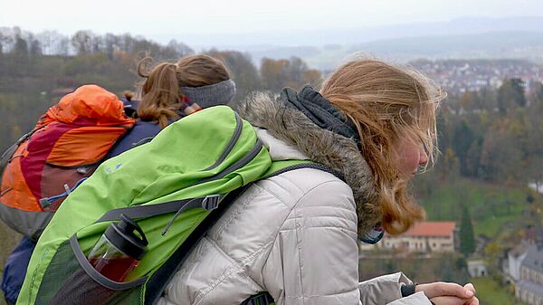 2 junge Frauen mit Rucksack sitzen vor Landschafts-Panorame